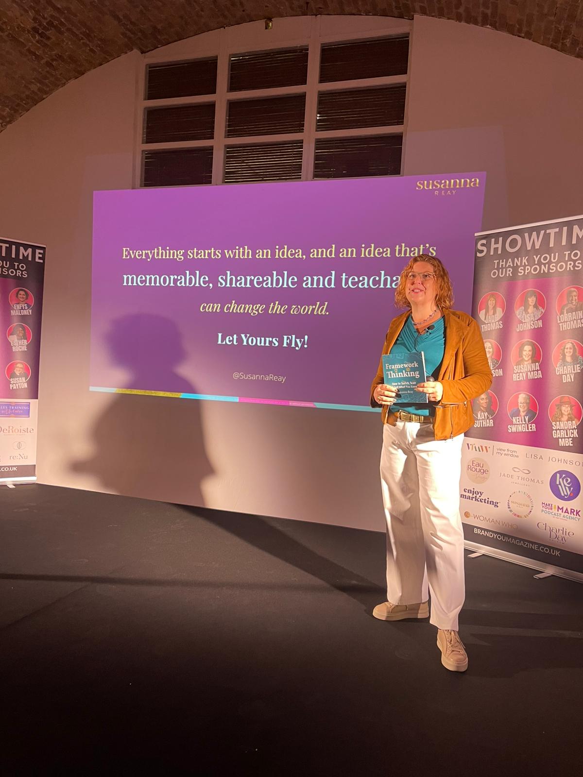 Susanna Reay speaking on stage at Showtime, Oct.2025 with a screen behind showing her big message "Everything starts with an idea, and an idea that is memorable, shareable and teachable, can change the world - so let yours fly". Susanna is holding a square turquoise book that is titled "Framework Thinking - How to sketch, scale and sell what you know"